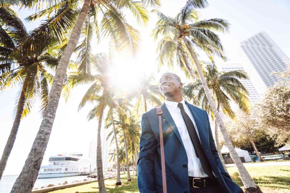 A smiling man in a suit near palm trees and a yacht in sunny Miami.